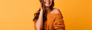 Adorable woman in orange attire touching her brown wavy hair. Laughing blithesome girl posing on yellow background.