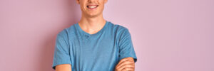 Teenager boy wearing casual t-shirt standing over blue isolated background happy face smiling with crossed arms looking at the camera. Positive person.