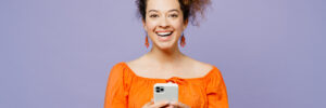 Young smiling happy cheerful latin woman she wear orange blouse casual clothes hold in hand use mobile cell phone isolated on plain pastel light purple background studio portrait. Lifestyle concept