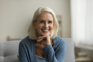 Cheerful pretty blonde senior woman looking at camera, smiling with healthy white teeth, laughing, posing for shooting on sofa, touching chin. Senior lady home head shot portrait